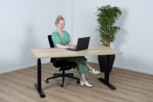 Vrouw kijkt naar haar laptop en zit achter een bureau met een licht eiken blad.