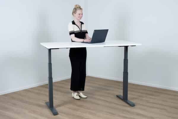 Vrouw staat achter een zit sta bureau met een antraciet kleurige poot en een wit bureaublad.