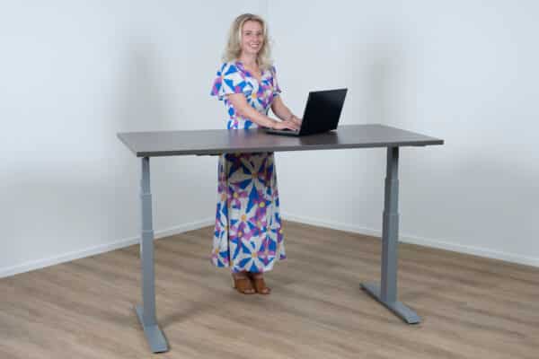 Vrouw staat achter een elektrisch bureau met een aluminium poot en een donker eiken blad.