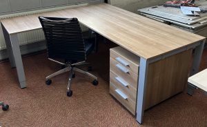 Tweedehands hoekbureau van het merk Vepa. Het heeft een donkereiken blad, met een ladekast eronder in dezelfde kleur. De poten en de grepen op het kastje zijn van aluminum. Achter het bureau staat een bureaustoel.