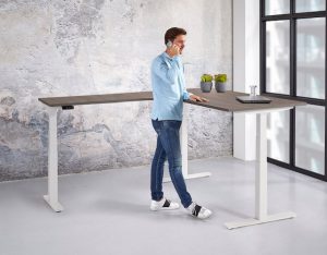 Kantoorruimte met daarin een hoekbureau. De aanbouw zit hier links. De poten zijn wit en een midden donker blad. Achter het bureau staat een man met lichtblauw overhemd.