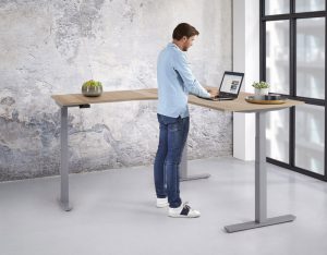 Een man werkt staand achter een hoekbureau met aluminium voet en een licht bureaublad.