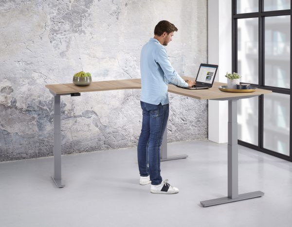Een man werkt staand achter een hoekbureau met aluminium voet en een licht bureaublad.