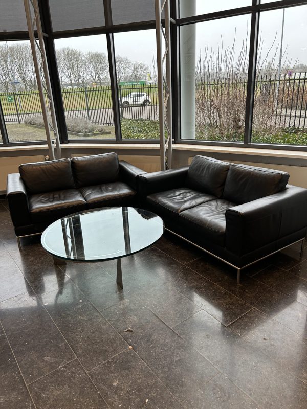 Ontvangsthal met daarin twee leren banken en daar tussenin een glazen salontafel. Elke bank is een gebruikt fauteuil in de kleur bruin, geschikt voor twee personen.