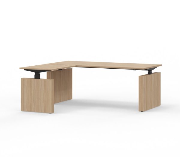 Witte achtergrond met daarvoor een L-bureau. Dit hoekbureau is van licht hout en heeft om de drie poten wangen. Deze is elektrisch verstelbaar.