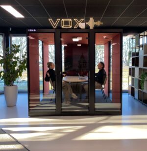 Zwarte vergadercabine met rode binnenkant, twee mensen overleggen erin.
