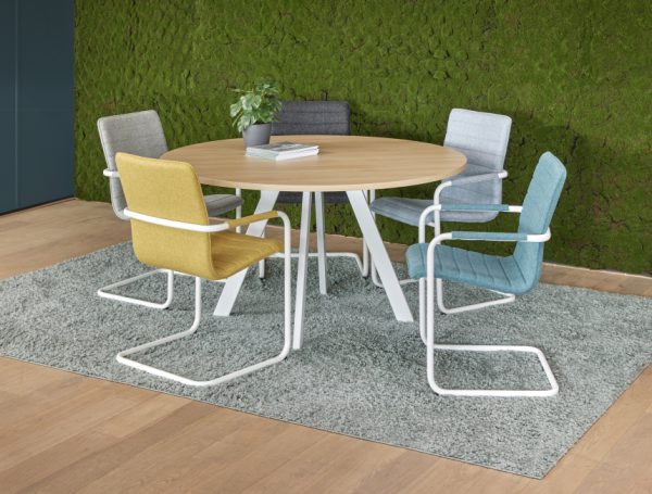 Kantoorruimte met daarin de vergadertafel rond met spinpoot. De poot is wit en het blad lichthout. Eromheen staan blauwe en gele kantinestoelen.