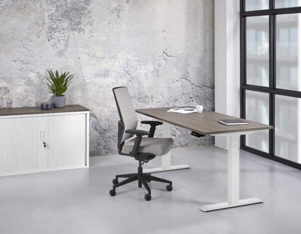 een grijze bureaustoel achter een zit sta bureau met witte poten en een bruin blad in een kamer met daarin een schuifdeurkast met daar bovenop een plantje Elektrisch zit sta bureau 140 x 80 professional