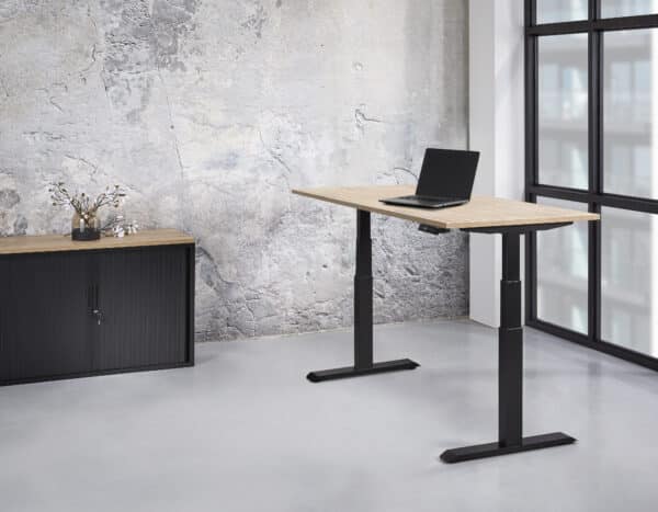 een zit sta bureau op zijn hoogste stand, met zwarte poten en een houten blad met daarop een laptop in een kamer met een schuifdeurkast Elektrisch zit sta bureau 140 x 80 professional