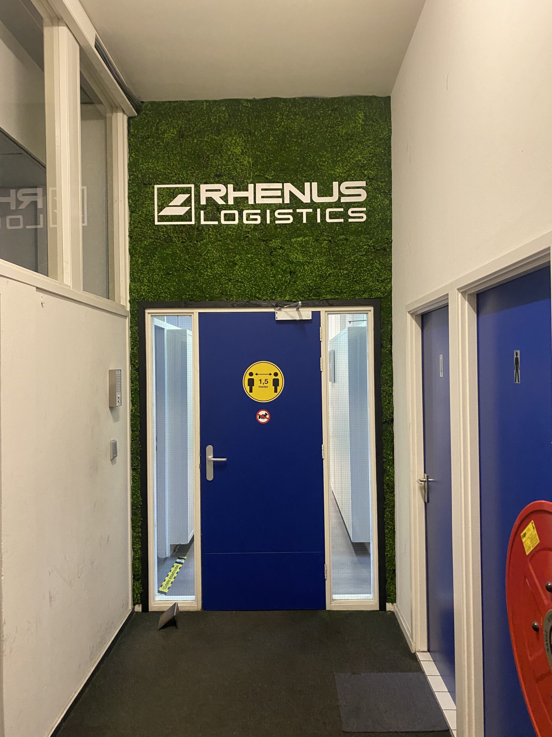 Gang met een deur aan het einde. Boven de deuropening zit een mos wand van kunstmos. Met daarop het logo van Rhenus Logistics.