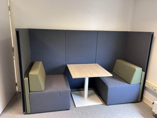 Kantoorruimte met daarin een zwart groene treincabine. De cabine bestaat uit zwarte akoestische panelen om de twee treinbanken heen. Tussen de twee treinbanken in staat een tafel. Dit is de Akoestische treinbank König + Neurath Gebruikt 02.