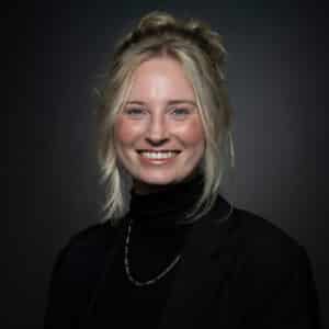 Portretfoto van een vrouw met blond haar in een knot en een zwarte trui en blazer aan.
