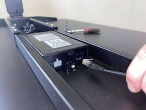 Hand steekt stekker van het bedieningspaneel in de controlbox van het elektrisch verstelbare bureau. 