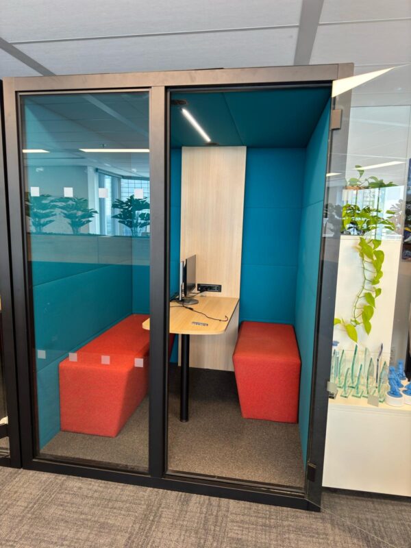 Tweedehands geluidsdichte cabine SIL05 met blauw interieur, licht houten tafel en rode bankjes erin. Glazen deuren voorin.