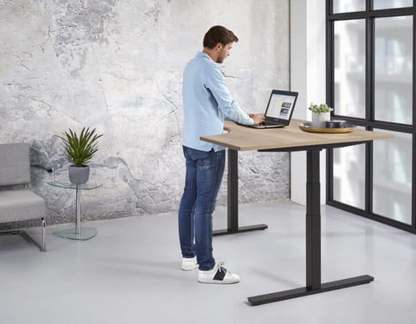 Man werkt staand achter een hoekbureau met zwart onderstel en licht houten blad.