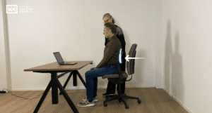 Screenshot van een zitinstructie met man op bureaustoel en docent.
